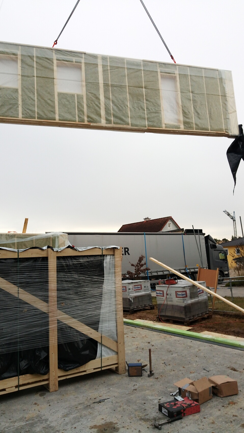 Große Wandbauelemente schwebend am Kran; Baustelle mit Paletten, Containern und Werkzeugen.