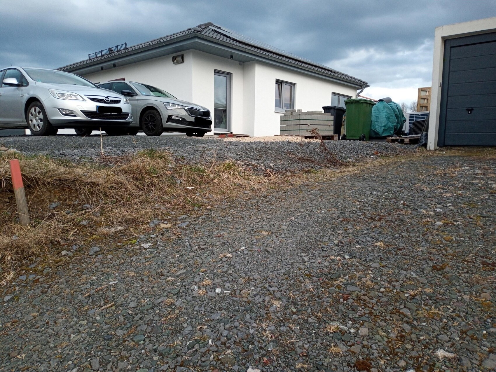 Einfamilienhaus mit Schotterhof, zwei Autos vorne, Mülltonnen und Paletten nahe Garage.