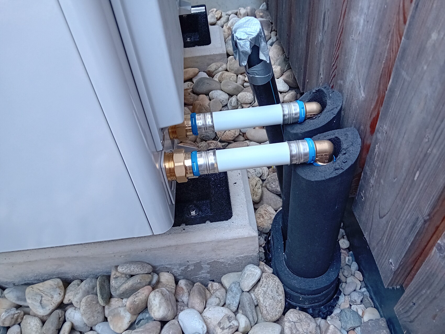 Außenanlage: Zwei isolierte Rohre mit Kupferfittings führen in ein Gerät; Kieselsteine und Holzzaun daneben.