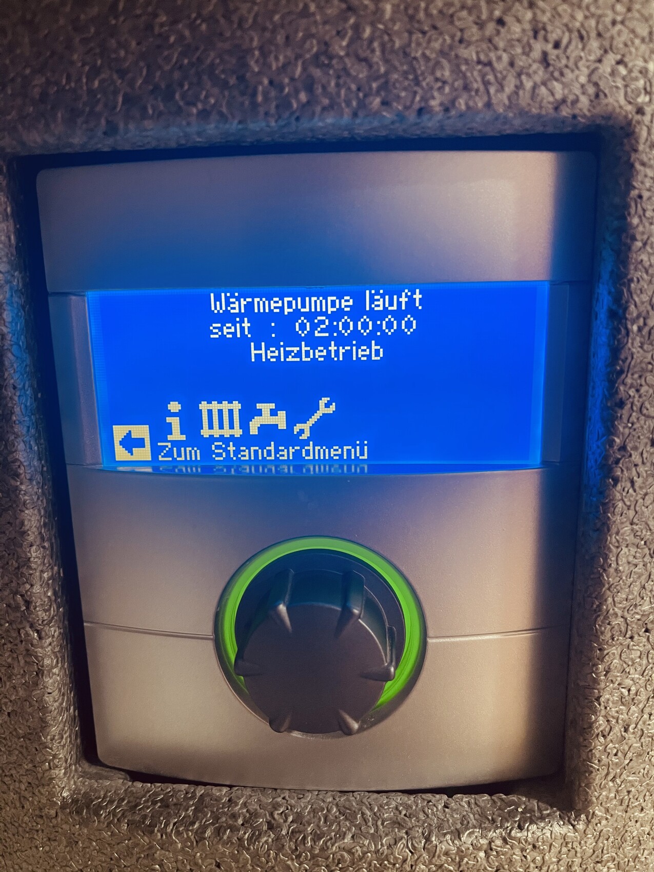 Blaues Display einer Wärmepumpen-Steuerung mit Laufzeit 02:00:00 und Heizbetrieb; grünem Rundknopf.