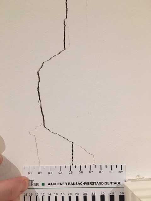 Riss in cremefarbener Wand, Messlineal mit Millimeter-Skala, Hand hält das Lineal.