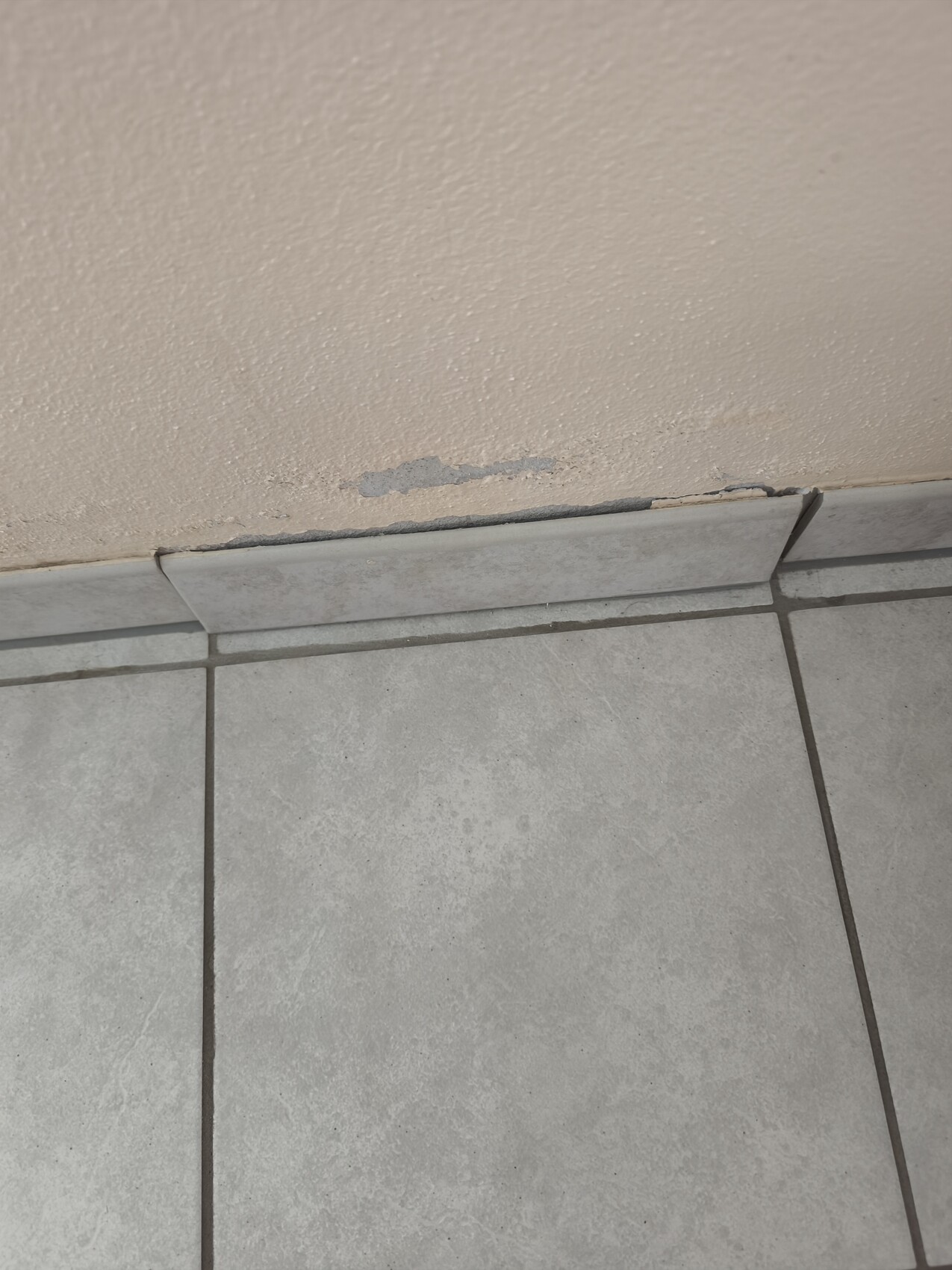 Beige Wand mit Abplatzungen an der unteren Kante über grauen Fliesen