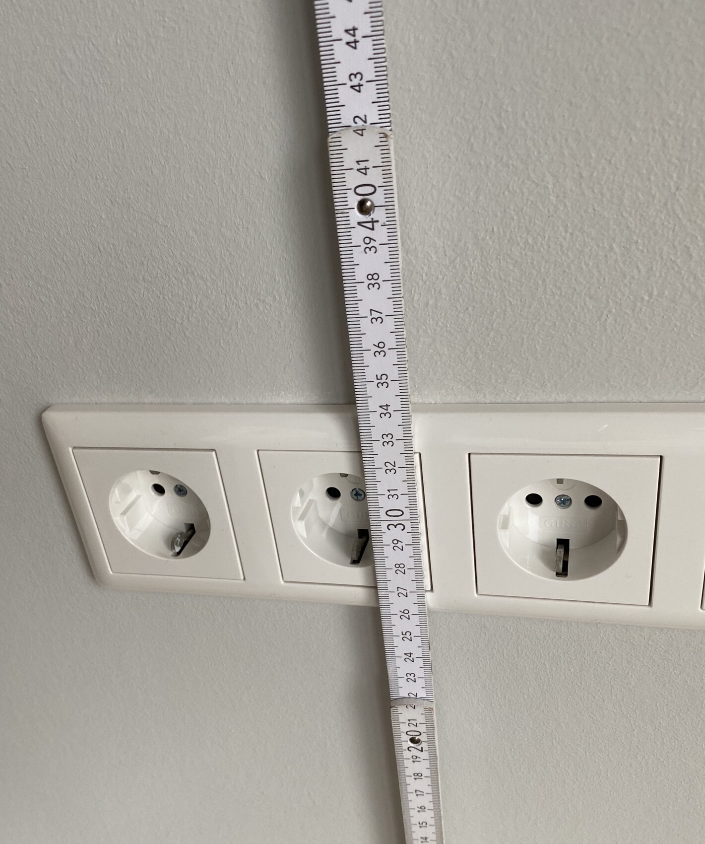 Maßband hängt senkrecht über drei Schuko-Steckdosen an der Wand.