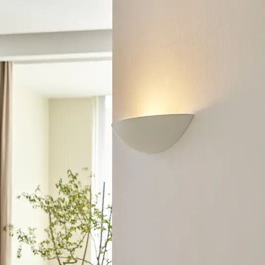 Heller Innenraum mit halbkugelförmiger Wandlampe rechts und Pflanze am Fenster links.