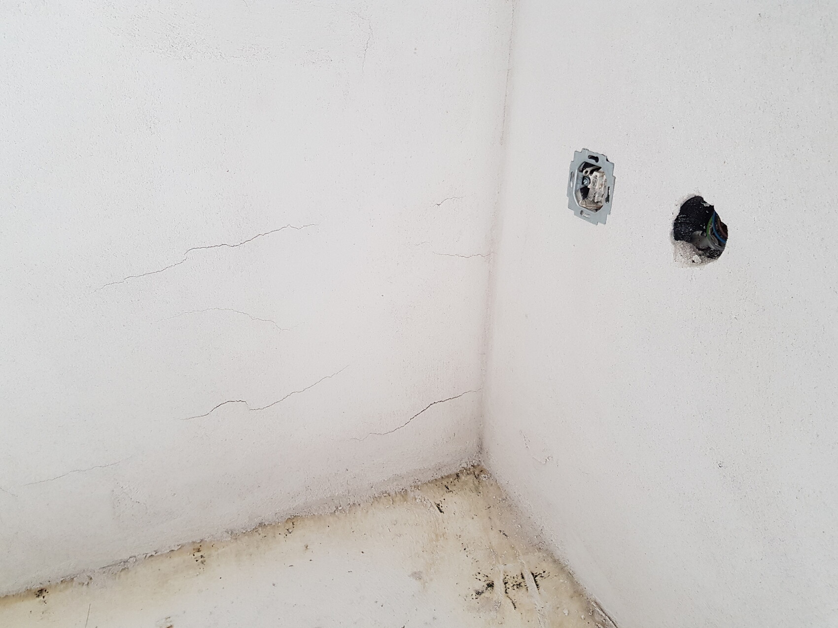Weiße Wand-Ecke mit Rissen; Steckdosen-Box und offenes Kabelloch, staubiger Boden.
