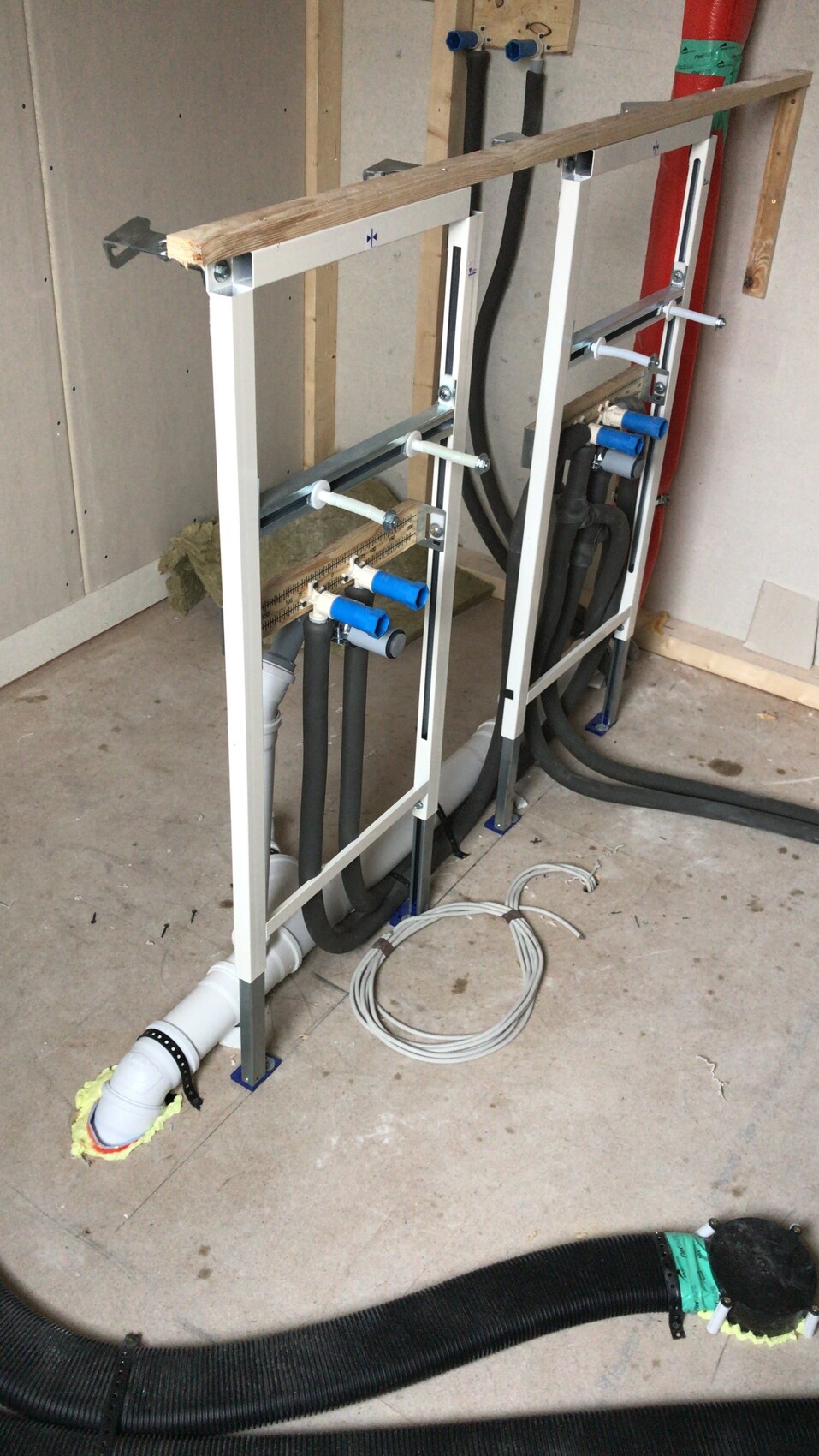 Zwei weiße Metallrahmen für Sanitärinstallation mit Rohren, Schläuchen und Kabeln in Bauumgebung.
