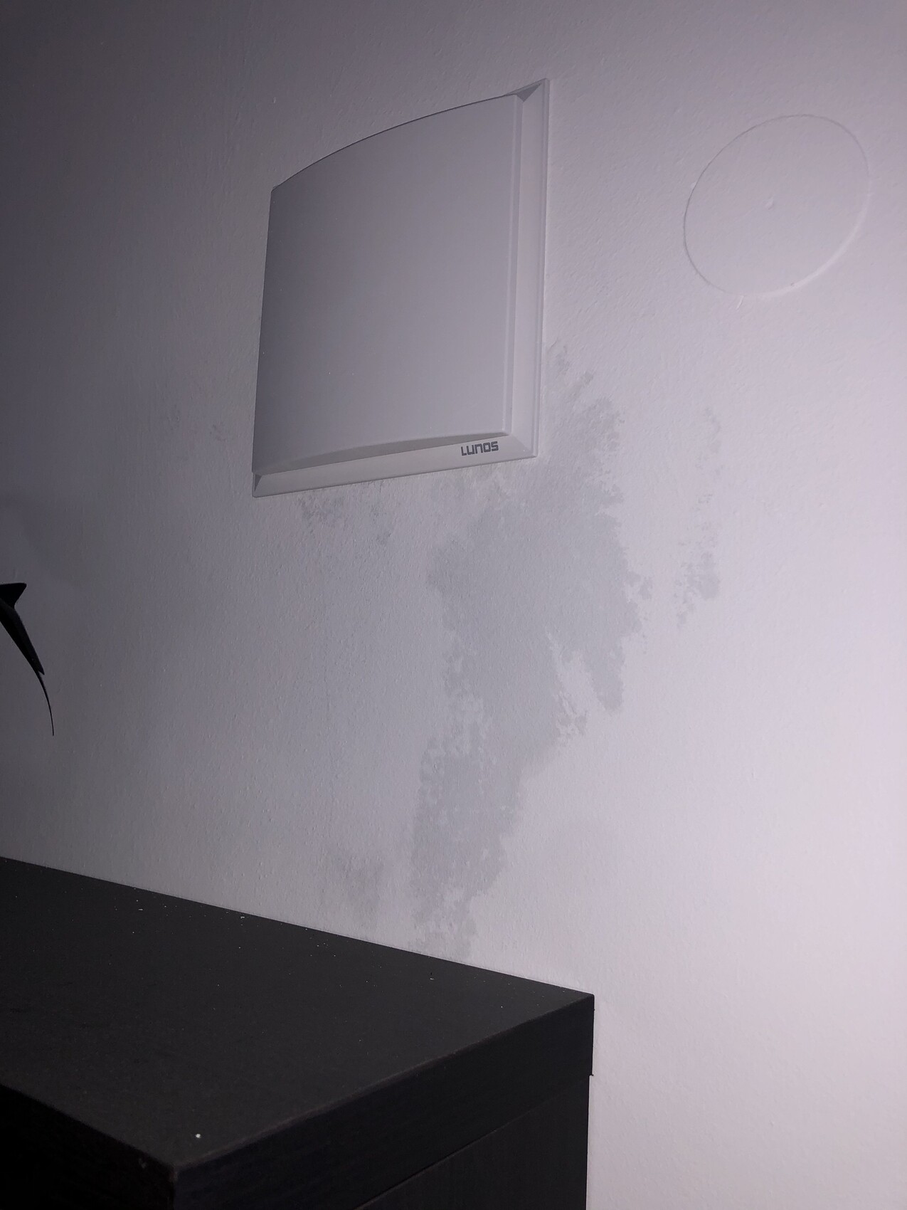 Weiße Innenwand mit Lunos-Wandlüfter über dunklem Schrank; sichtbare Feuchtigkeitsflecken.