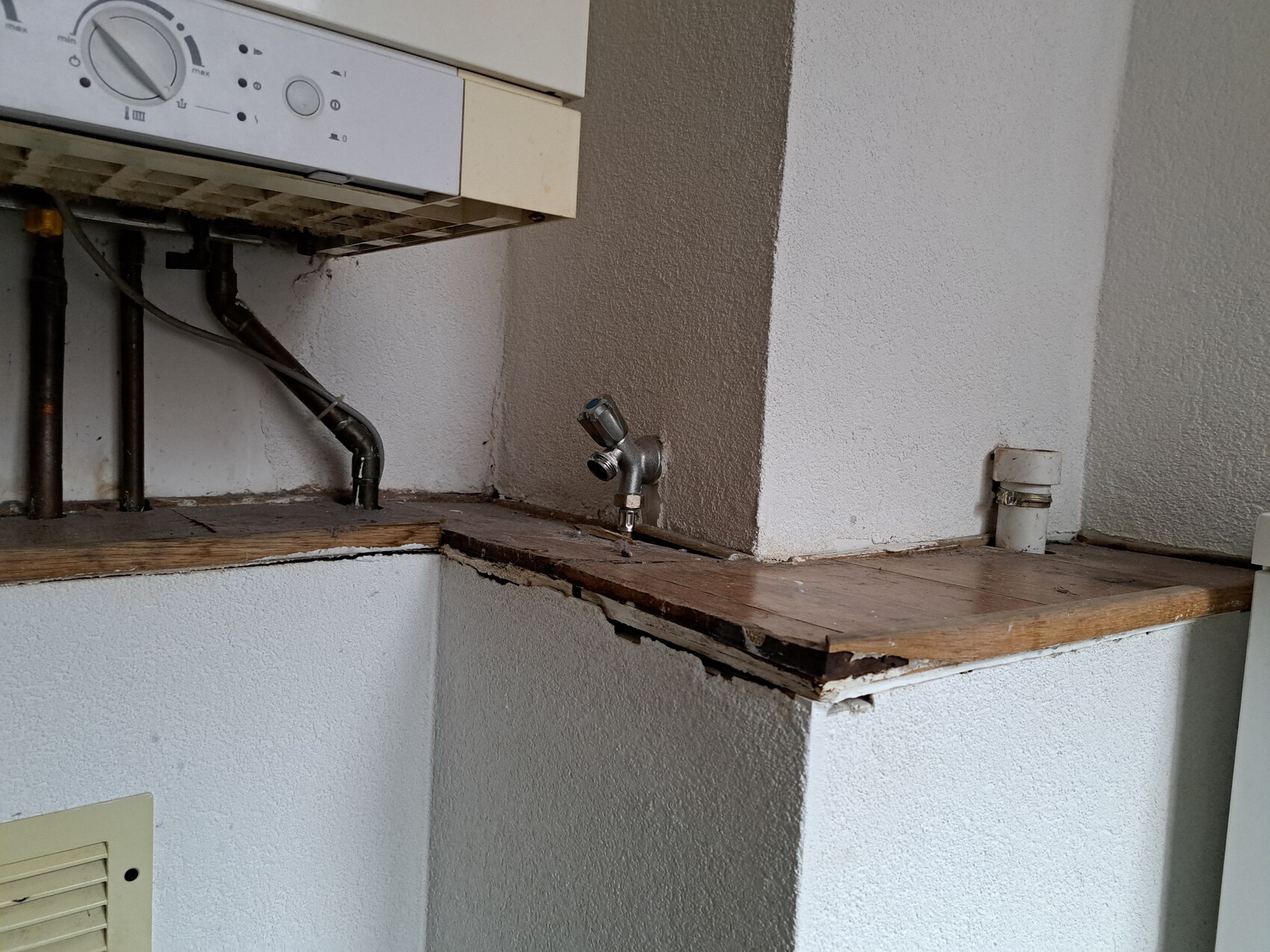 Waschmaschine mit Rohrleitungen darunter; Wasserhahn auf Holzplatte, beschädigte Kante.