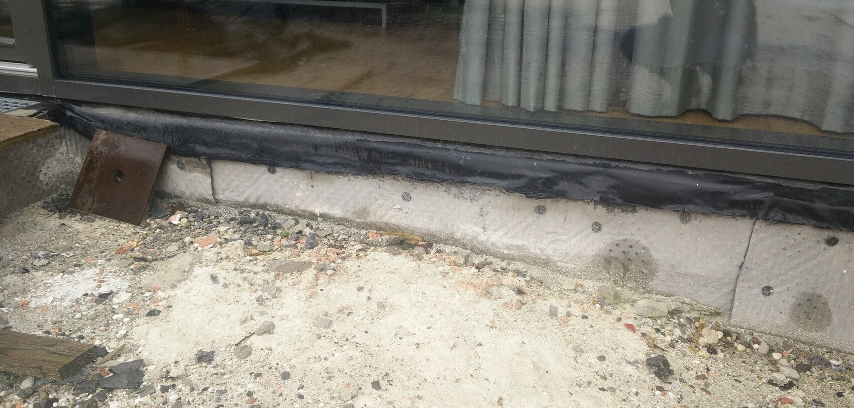 Außenansicht eines Gebäudes: Fenster mit schwarzer Abdichtung am Sockel, Schutt und Metallplatte davor.