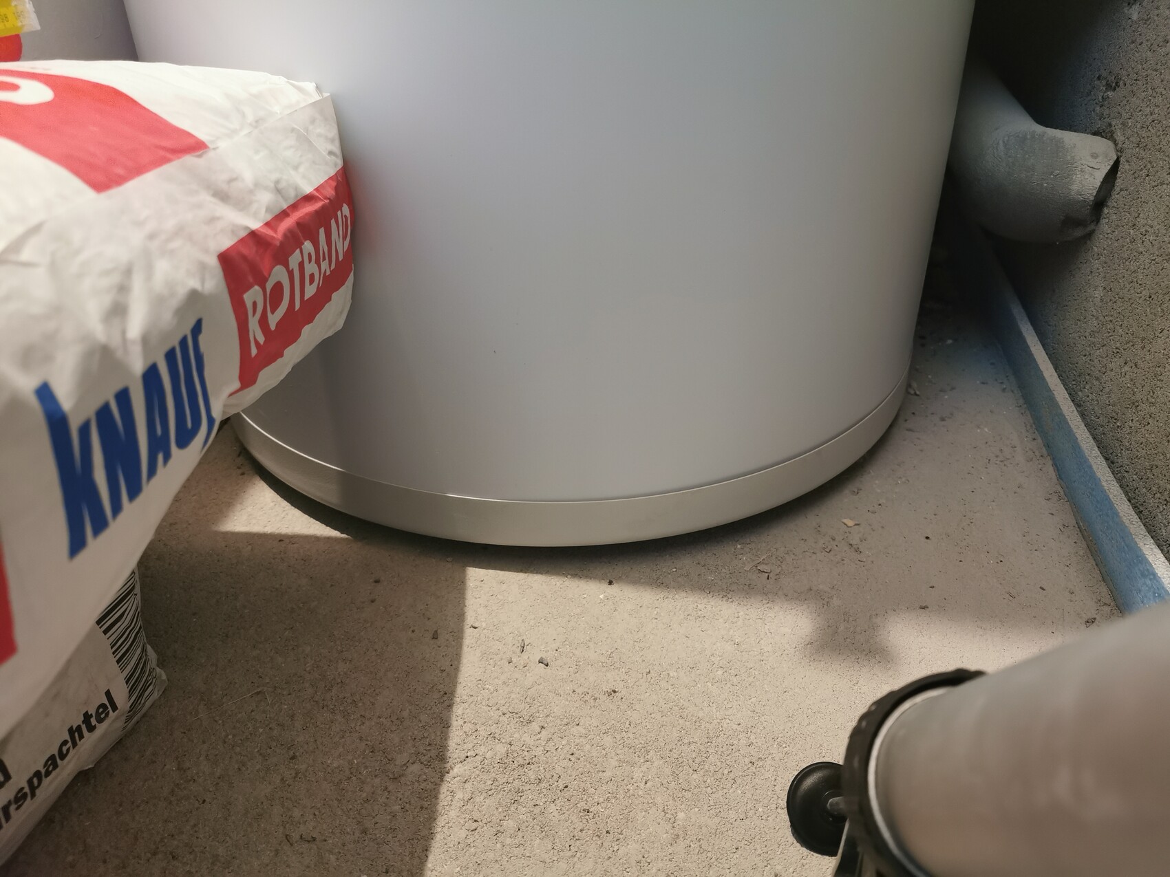 Großer weißer Zylinder-Boiler steht auf staubigem Boden neben einem Rotband-Zementsack im Keller.