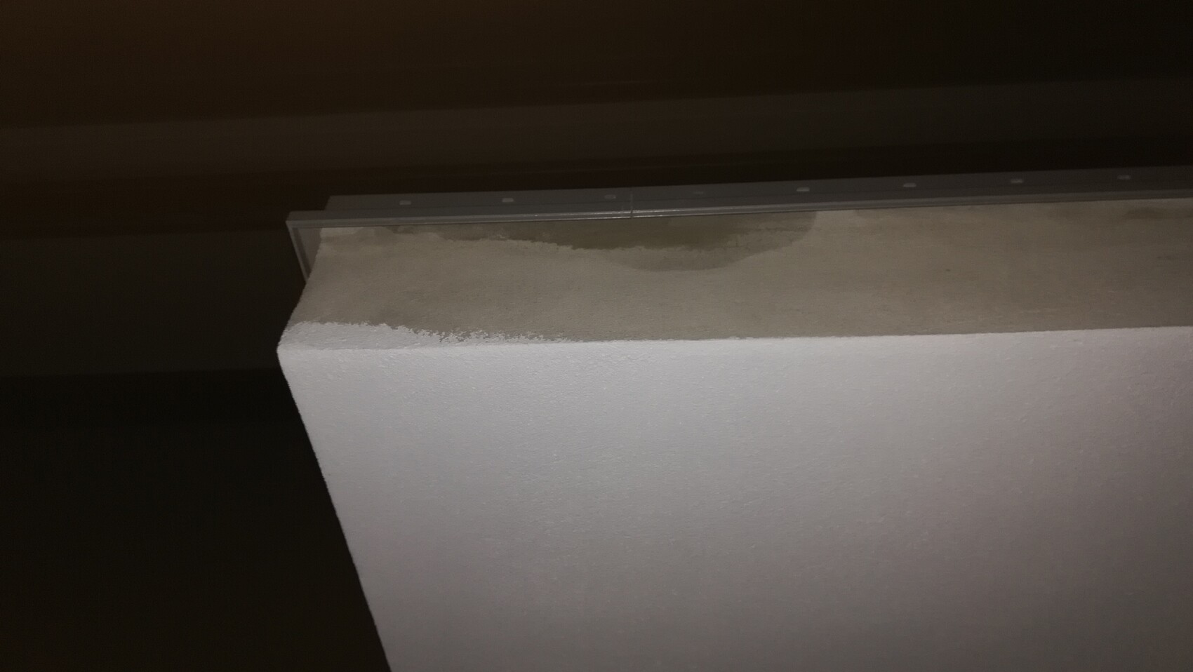 Nahaufnahme einer weißen, rauen Wand mit beige-grauer Oberseite und Metallleiste am oberen Rand.