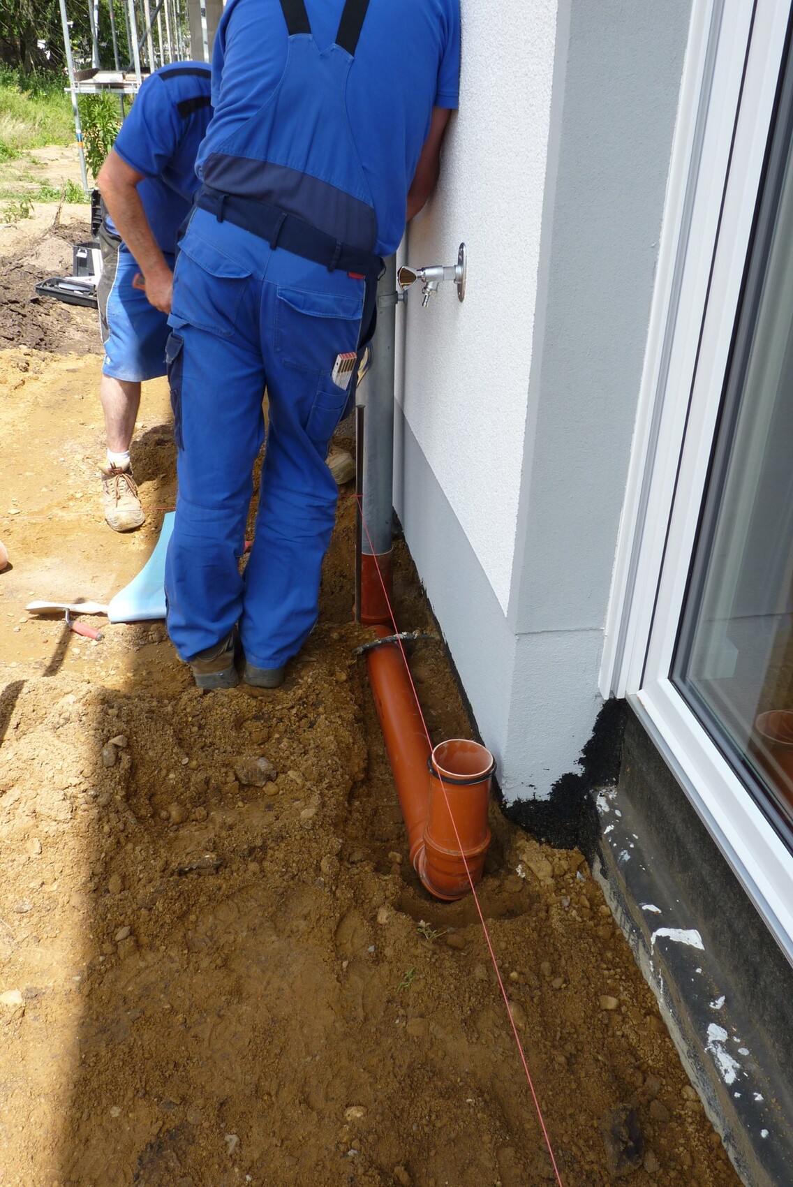 Bauarbeiter installieren eine orange Wasserleitung außerhalb eines Hauses am Fundament.