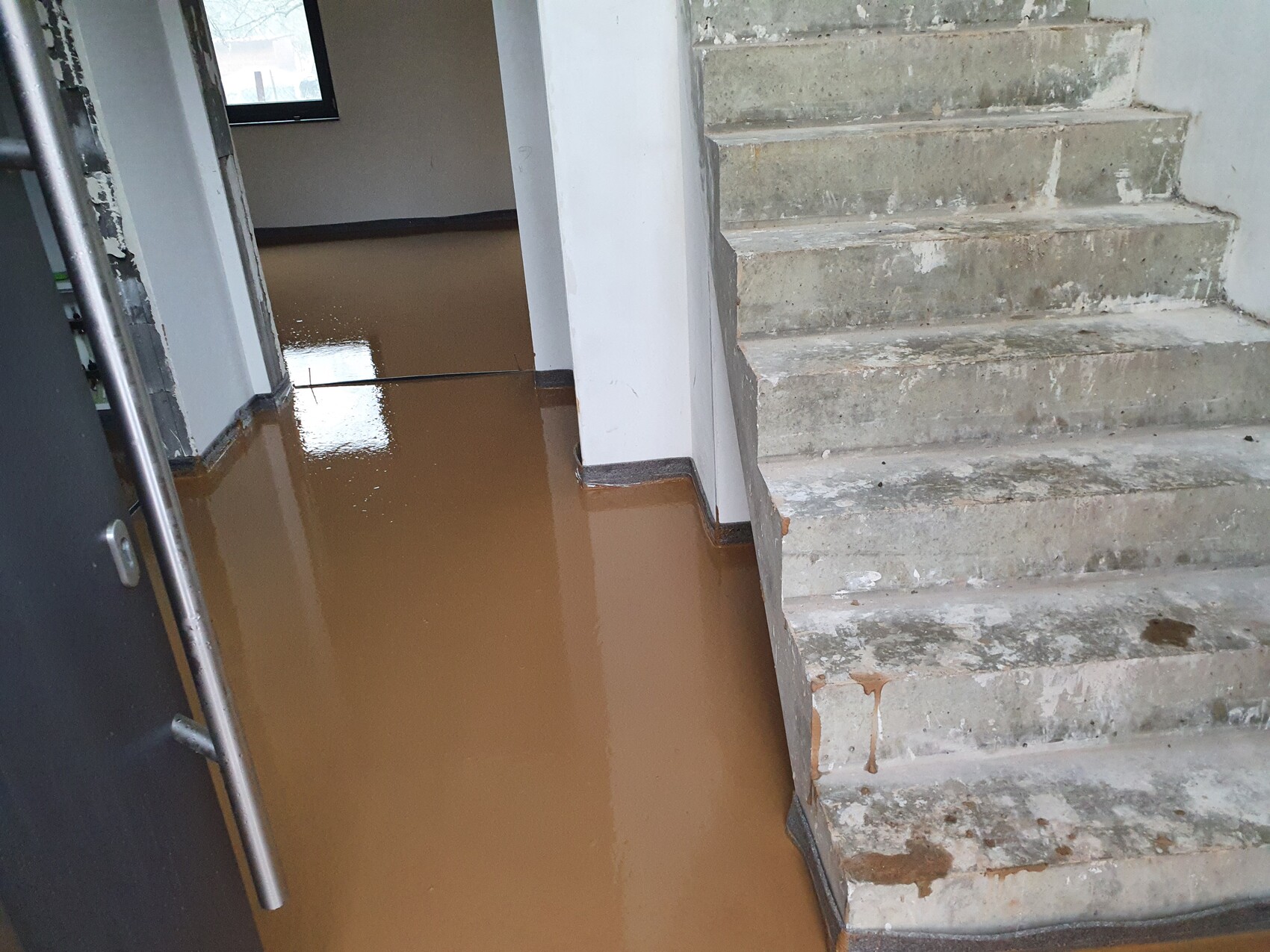 Überfluteter Innenraum mit braunem Wasser auf dem Boden; linke Türöffnung, rechte Treppe aus Beton.