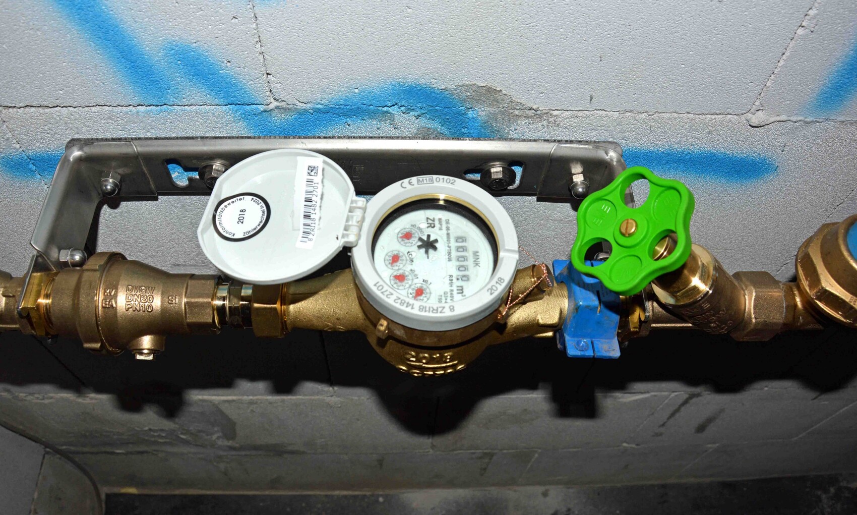 Wasserzähler und Rohrleitung mit grünem Absperrhahn vor grauer Wand mit blauem Graffiti.