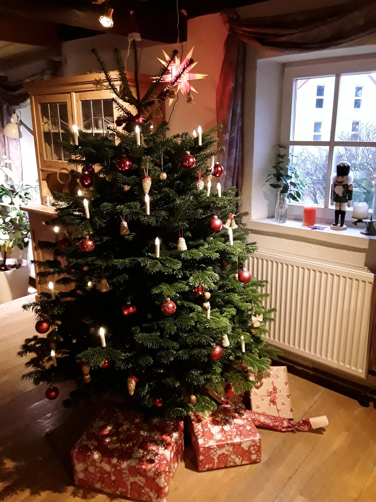 Weihnachtsbaum im Wohnzimmer mit Kerzen, roten Kugeln, Geschenken; Nussknacker auf Fensterbank.