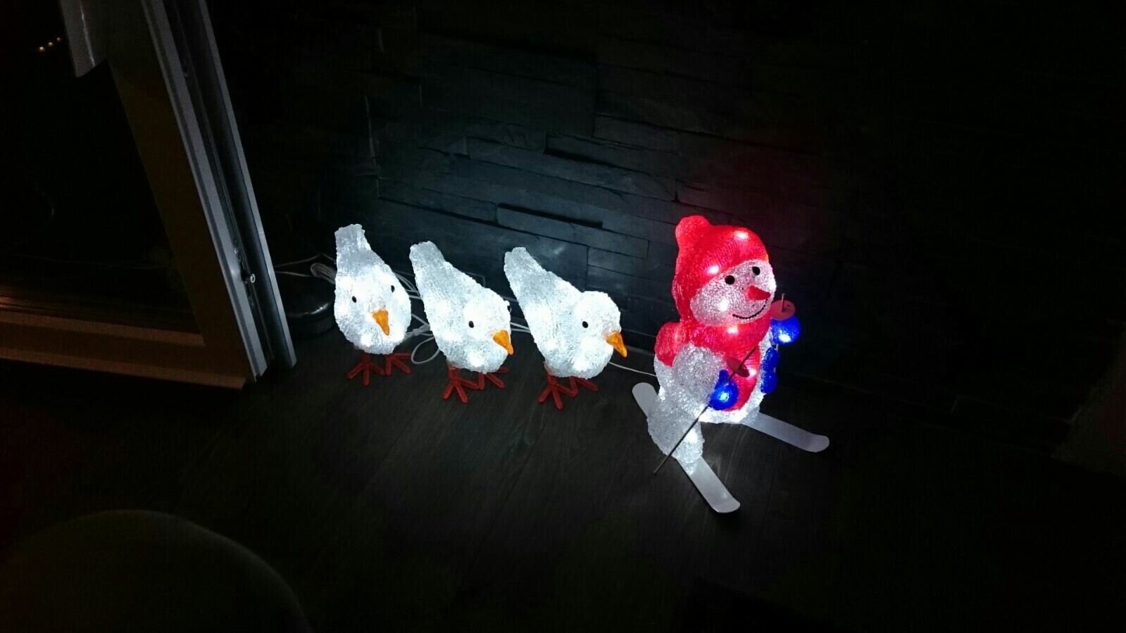 Leuchtende Weihnachtsdeko: drei weiße Vögel mit orangen Schnäbel folgen rotem Schneemann auf Skiern.