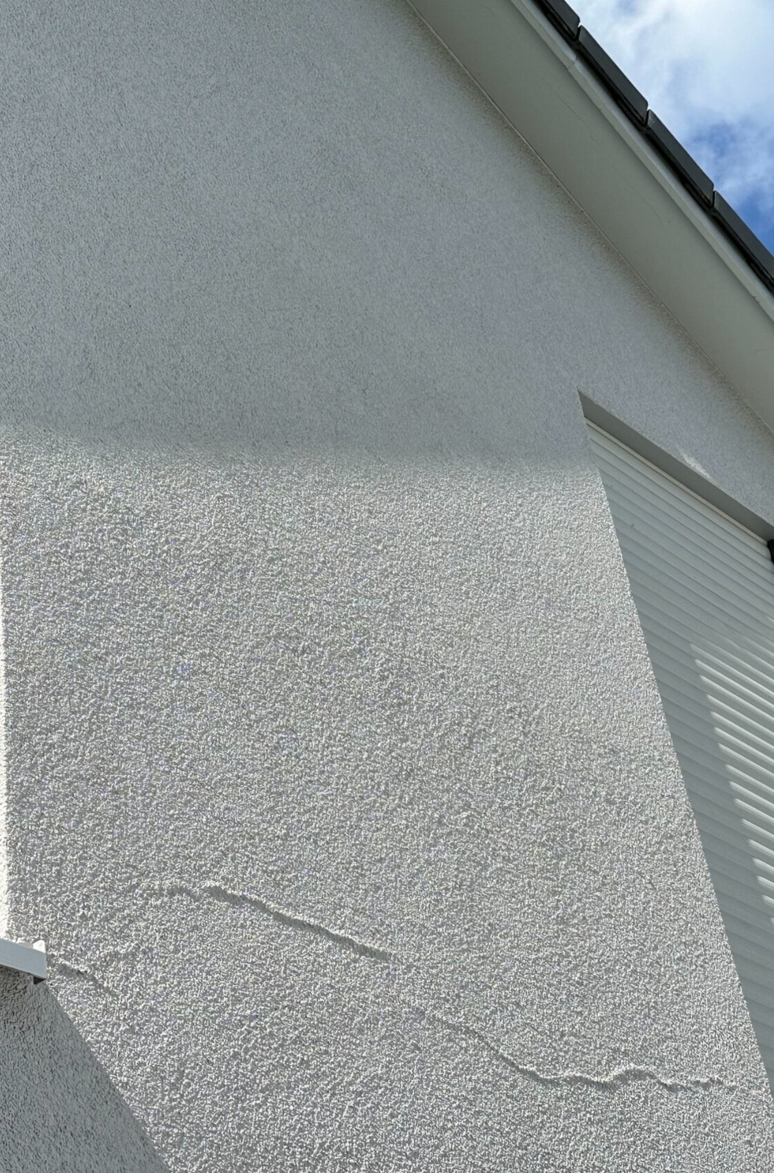 Weiße grob strukturierte Außenwand mit feinen Rissen; rechts Rollladenkasten, blauer Himmel