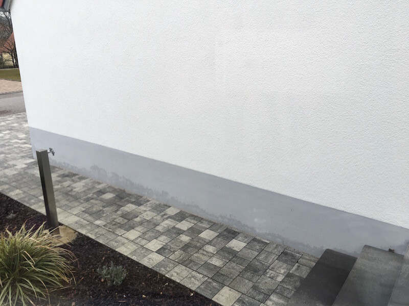 Weiße Außenwand mit grauer Sockelzone, gepflasterter Weg, Stufen rechts und Pflanzen links.