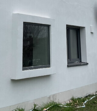 Weiße Außenwand eines Gebäudes mit zwei grauen Fensterrahmen; Frontfassade und Grasboden.