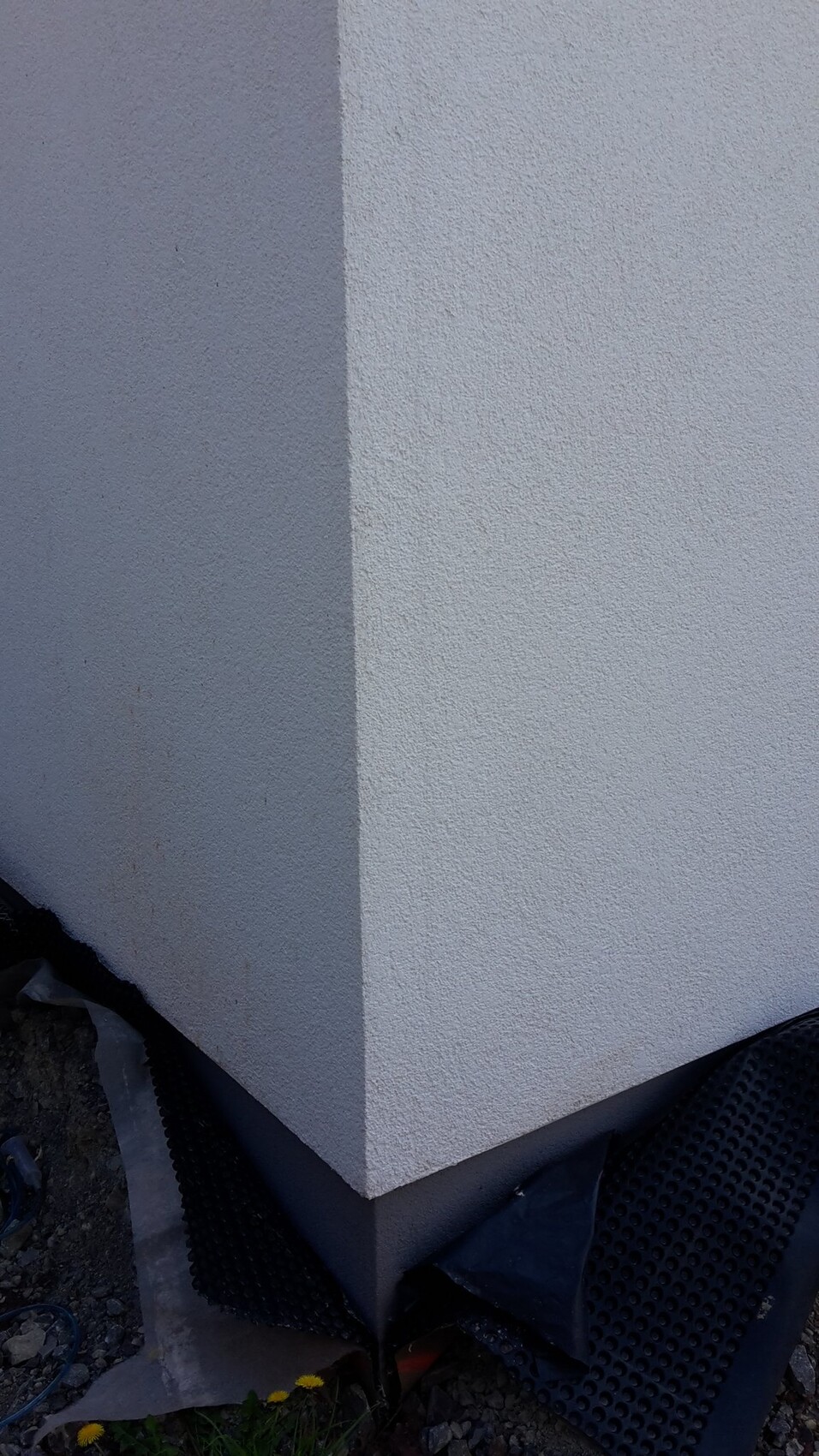 Ecke einer weißgrauen, rauen Außenwand mit dunklem Sockel; Boden mit Bauabfällen.