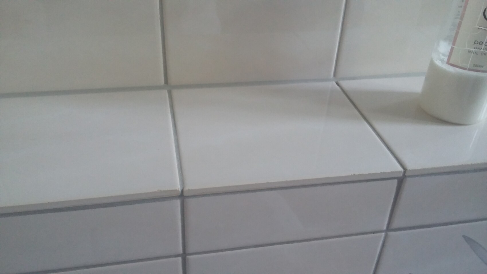 Weiße quadratische Fliesenwand; rechts steht eine weiße Flasche auf der Fliesenoberfläche.