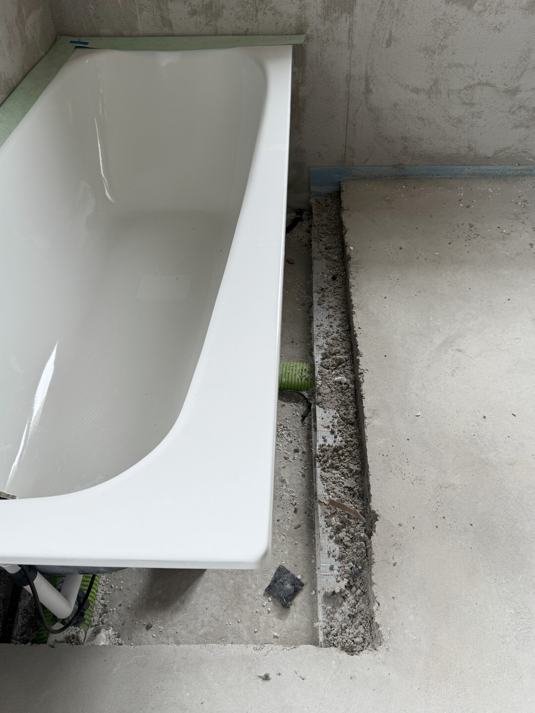 Weißer Badewannenrand im Rohbau, freigelegter Bodenkanal mit Schutt und grüner Leitung sichtbar