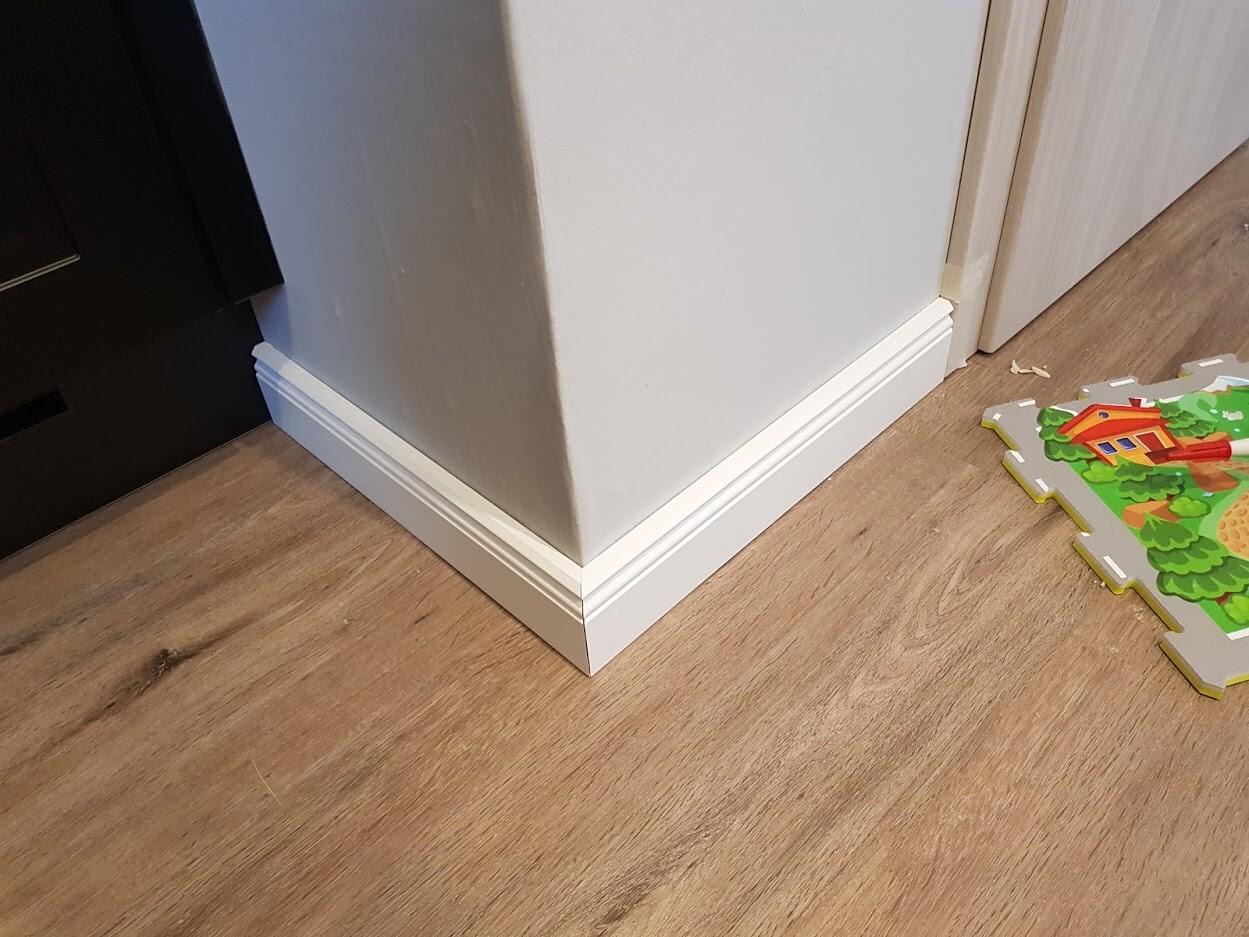Eckansicht eines Zimmers: weiße Sockelleiste, Holzfußboden, bunte Spielmatte rechts.