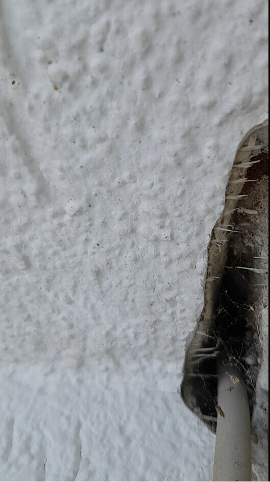 Nahaufnahme einer weißen, rauen Wand mit Loch, durch das ein Rohr sichtbar ist.