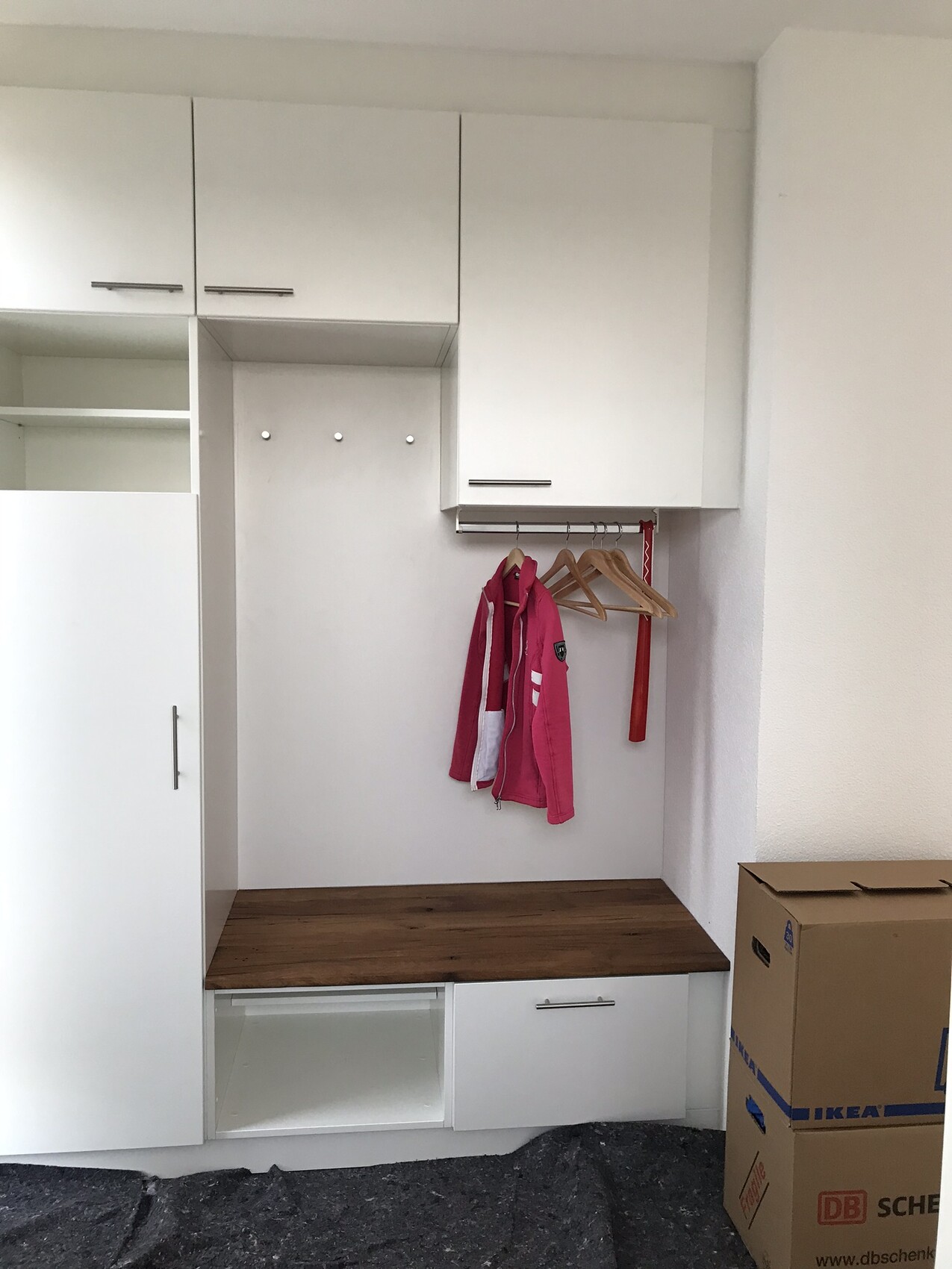 Weiße Einbau-Garderobe mit Holzplatte, Hakenleiste und pinker Jacke; Umzugskartons rechts.