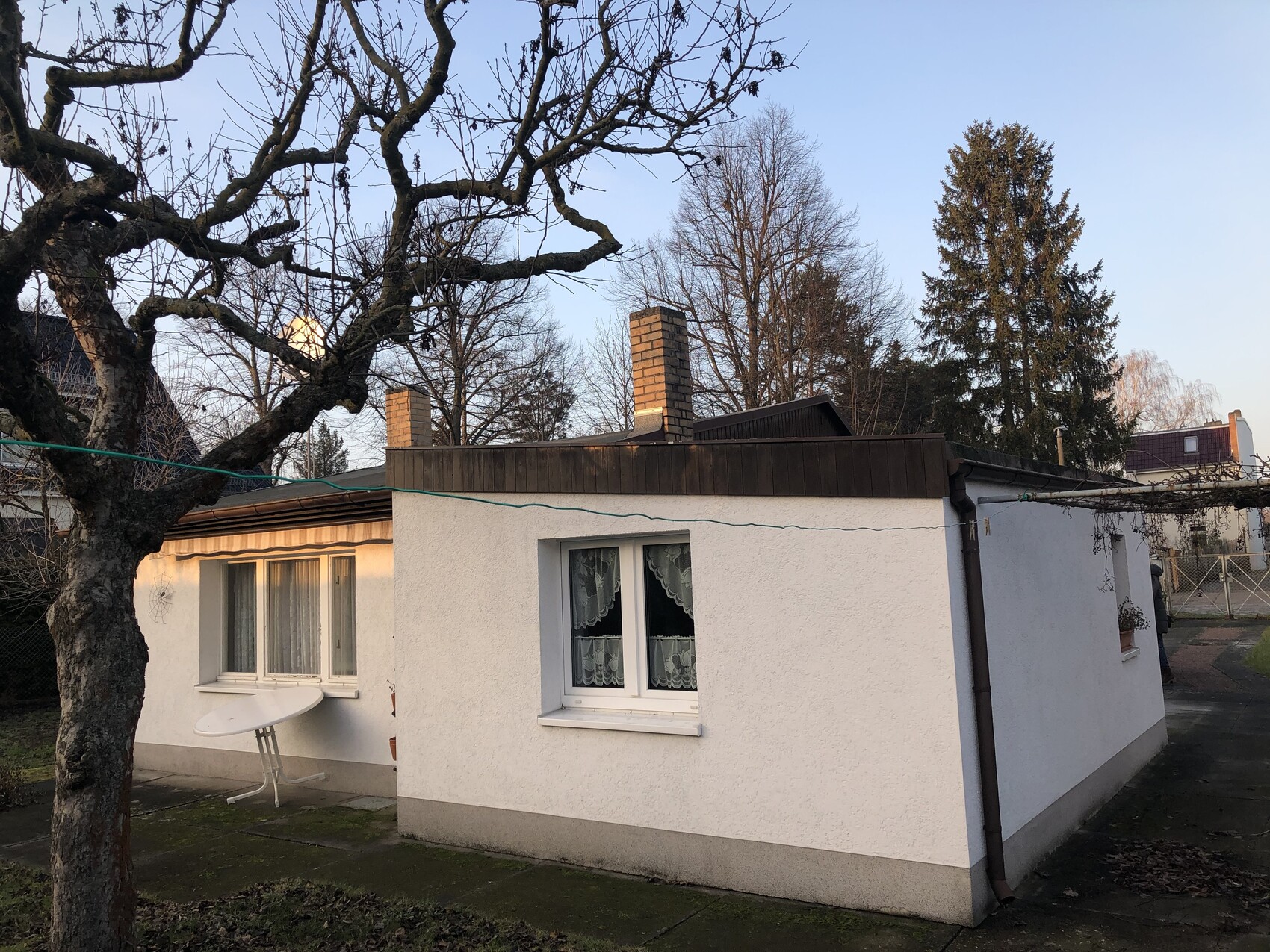 Kleines weißes Einfamilienhaus mit Flachdach, Kamin; kahler Baum im Vordergrund, Fenster mit Vorhängen.