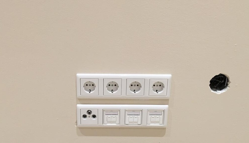 Wand mit vier oberen Schuko-Steckdosen, unten weitere Steckdosen; Loch in der Wand rechts.