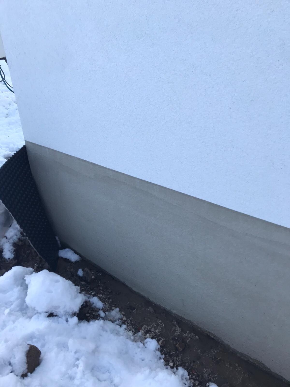 Außenwand mit weißem Putz, grauem Fundament, Schnee am Boden.