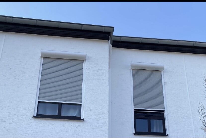 Weiße Hauswand mit zwei Fenstern, heruntergelassene Rollläden, blauer Himmel