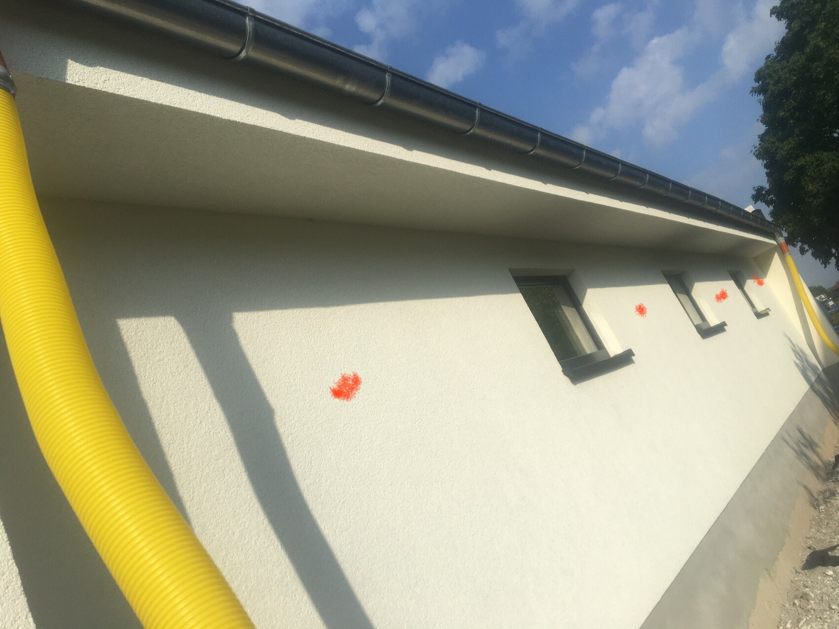 Weiße Gebäudewand mit gelben Rohren, orange Flecken und schmalen Fenstern.