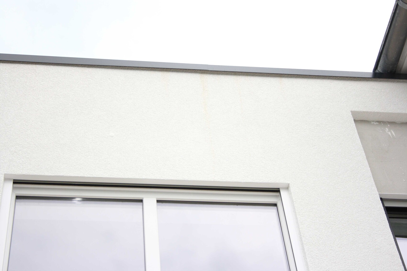 Weiße verputzte Außenwand mit Fenster unten links; oben graue Kante, leichte Wasserflecken.