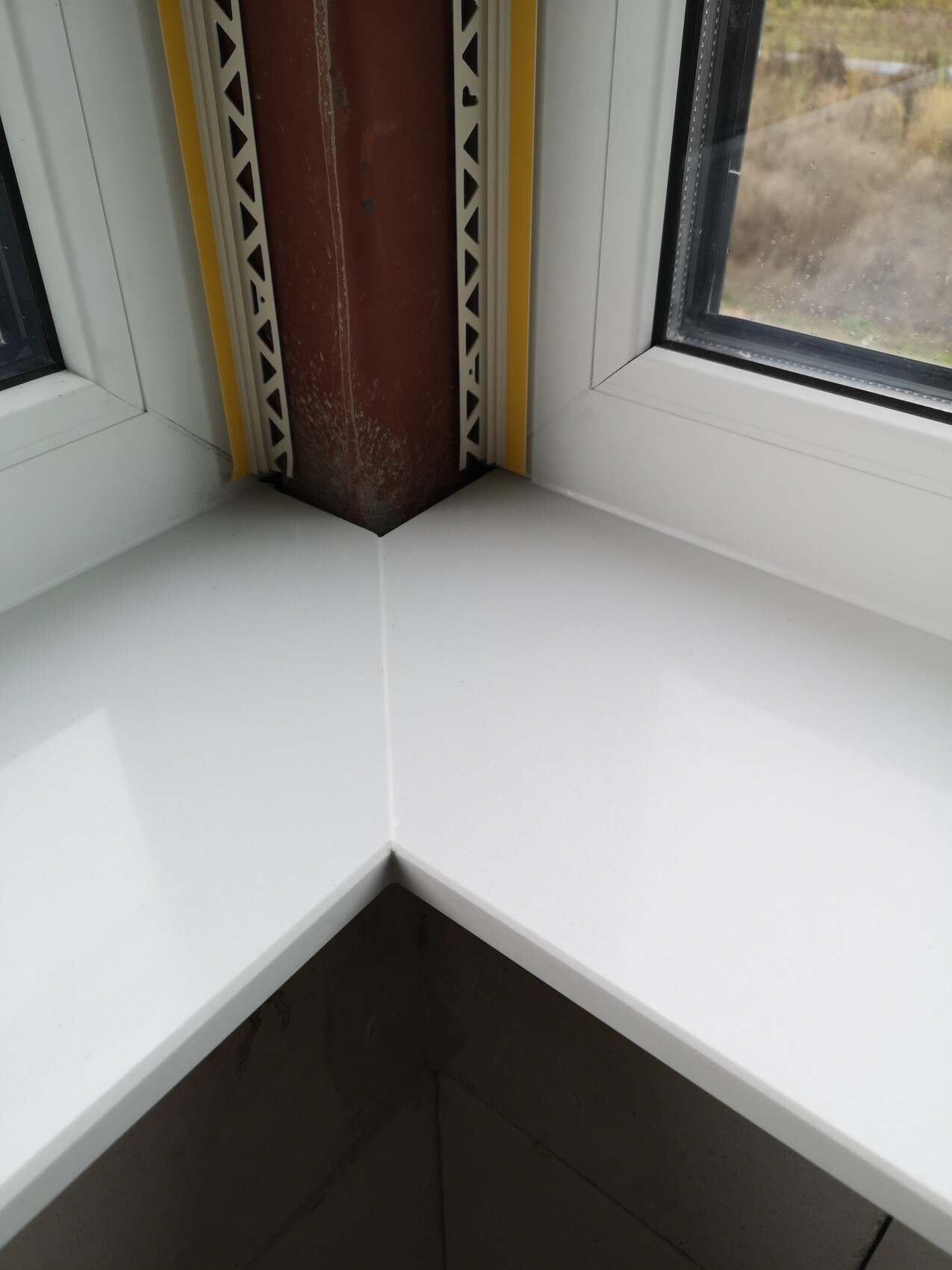 Weiße Fensterbank-Ecke mit braunem Pfeiler, gelbem Rand und Lochblechleisten.