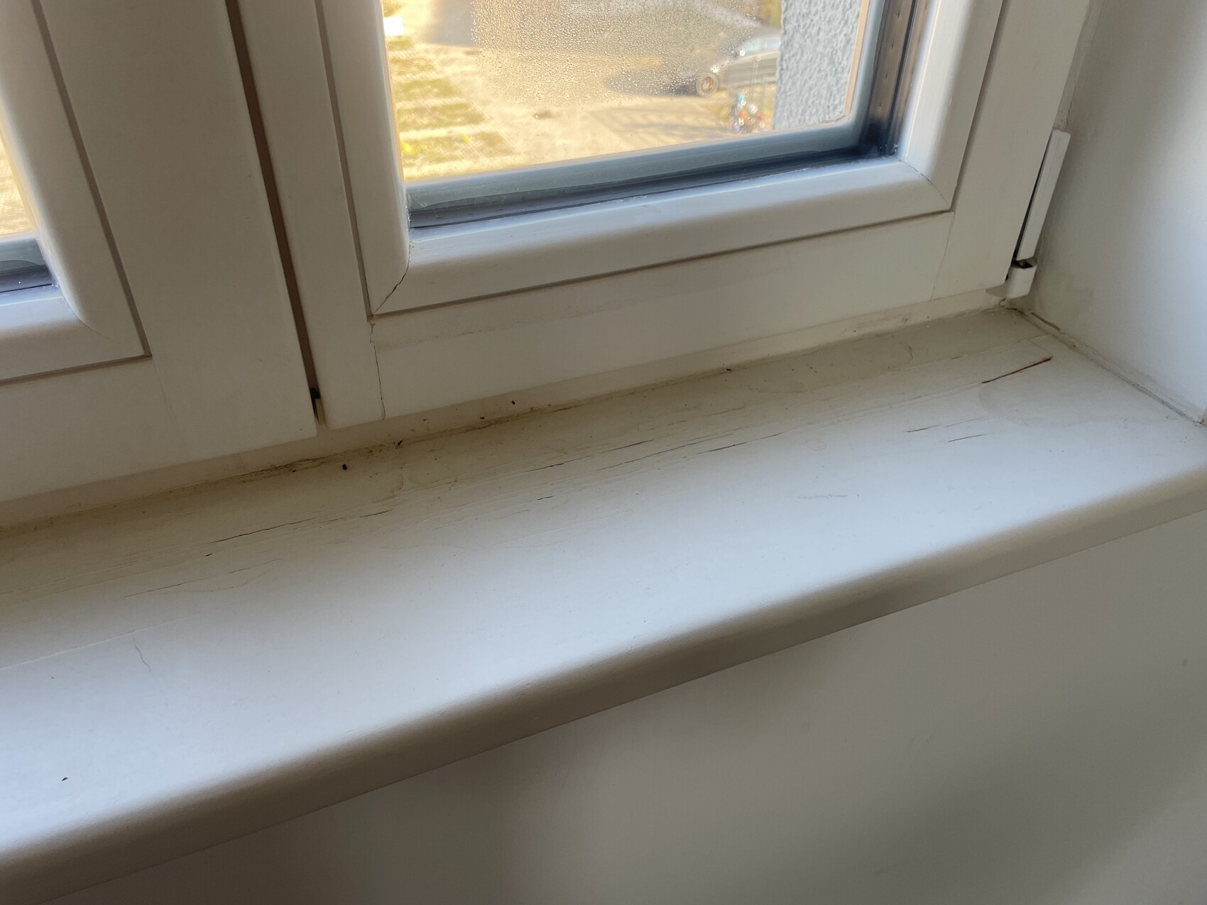 Weiße Fensterbank mit Staub und feinen Rissen; Blick durch Fenster auf Parkplatz und Auto.