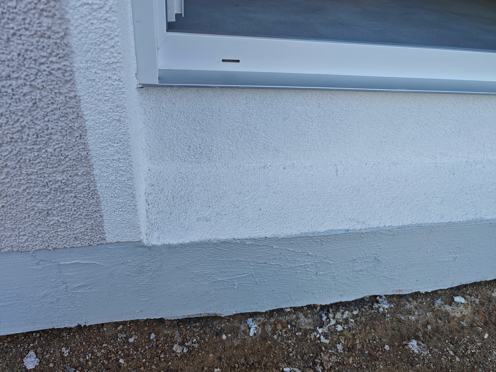 Außenwand mit Fenster: weiße Putzfassade, strukturierte linke Fläche, grauer Betonsockel, Erdreich unten.