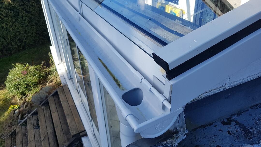 Weiße Dachrinne mit Fallrohr an Glasveranda, nahe grünem Garten.