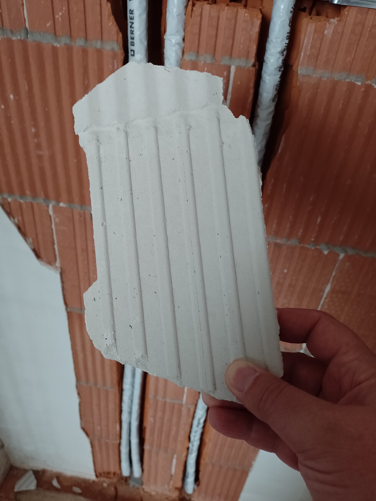 Eine Hand hält ein weißes Plattenfragment mit vertikalen Rillen vor einer Ziegelwand mit Rohren.