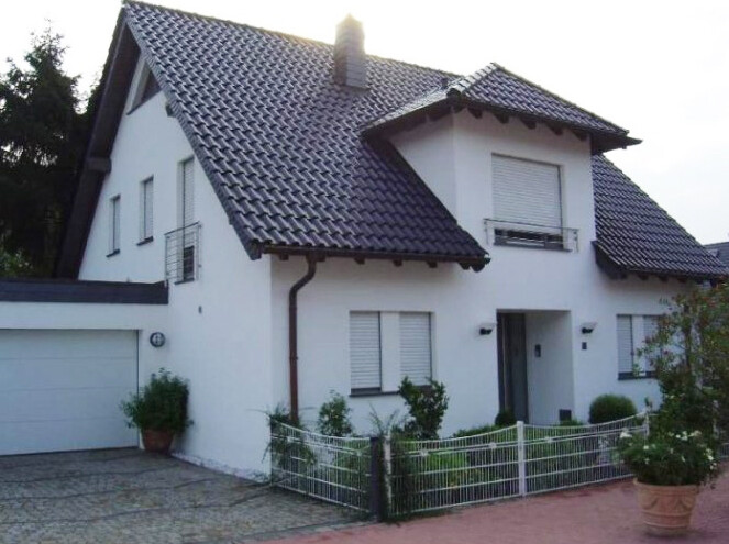 Weißes, zweigeschossiges Haus mit dunklem Ziegeldach, Garage und Garten hinter Zaun.