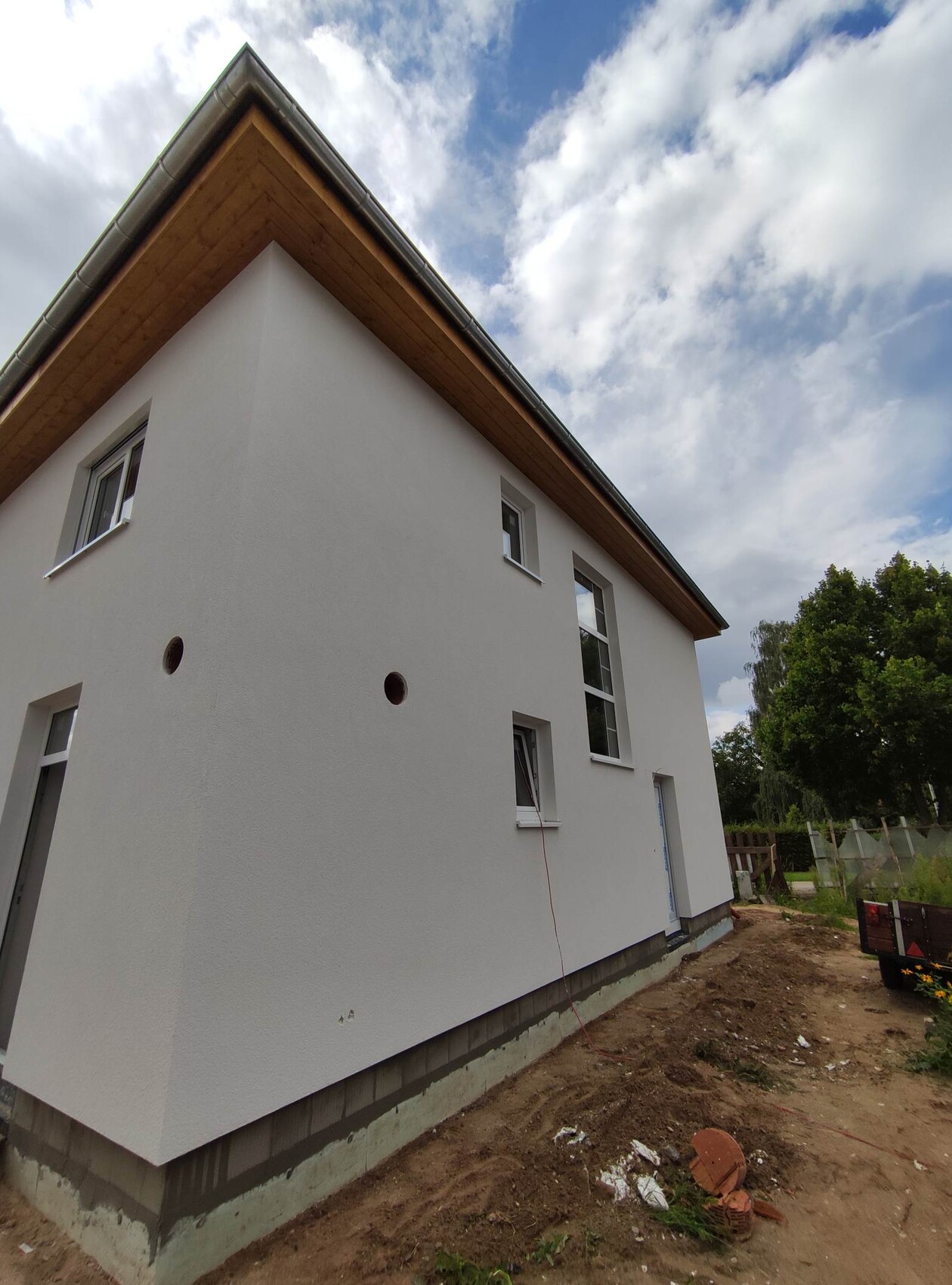 Außenansicht eines weißen Hauses im Bau mit Holzdachüberstand, Fenstern und Erdarbeiten.
