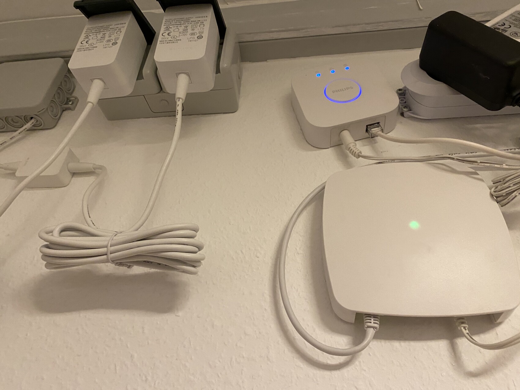 Weißer Tisch mit mehreren Netzteilen, Kabeln und zwei weißen Geräten; blau leuchtende LED-Anzeige.