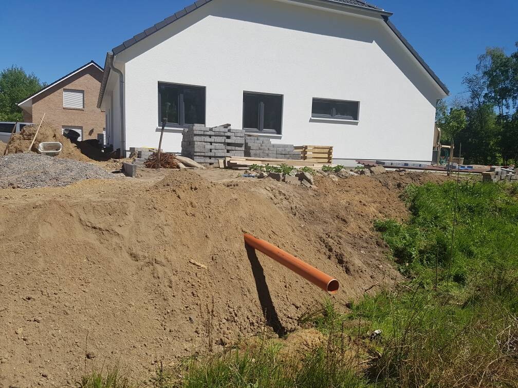 Weißes Einfamilienhaus im Bau, Erdaushub vorne, orangenes Rohr aus Graben, Baustoffe daneben