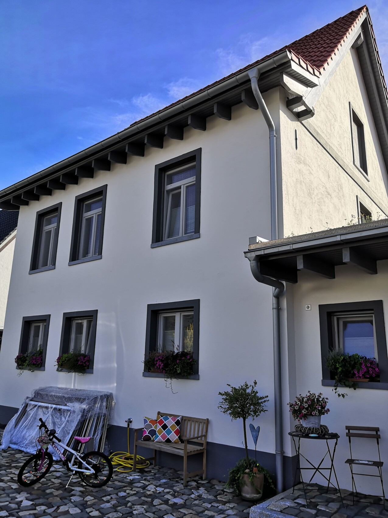 Zweistöckiges weißes Haus mit grauen Fensterrahmen, Blumenkästen, Hof mit Fahrrad.