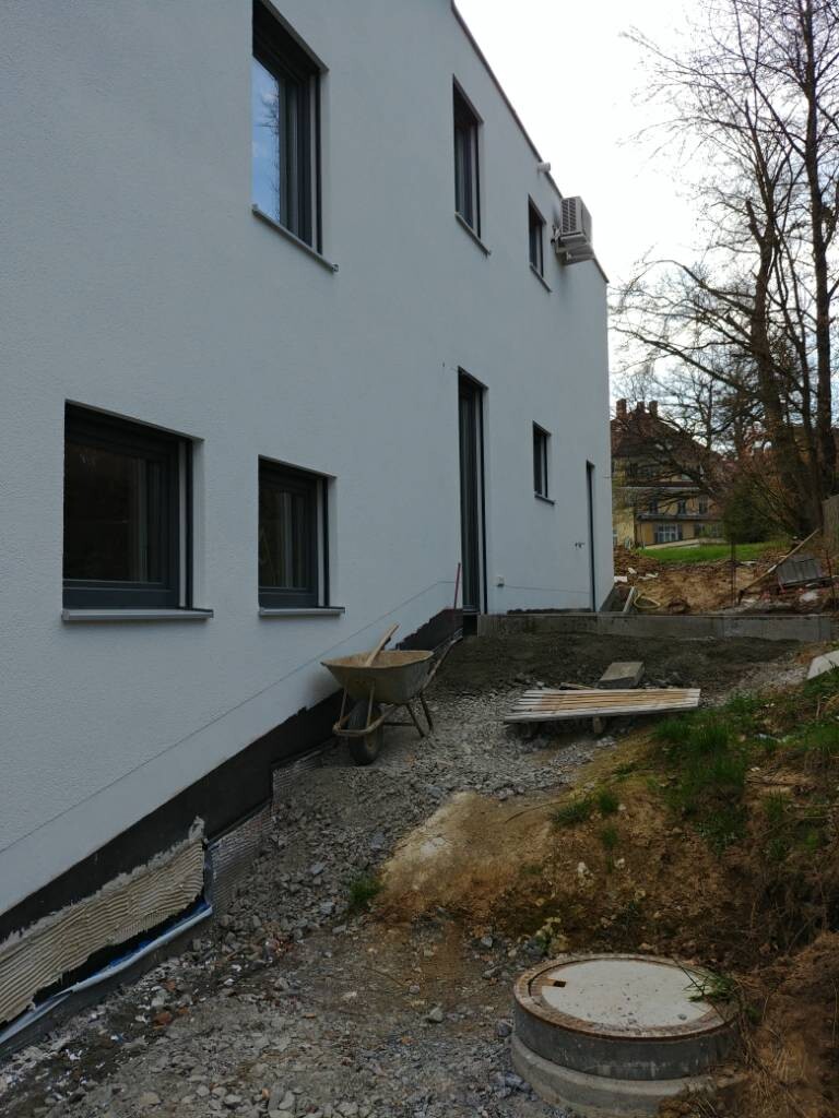 Weiße Gebäudefassade mit Fenstern; Bauarbeiten außen, Schubkarren, Holzplanken und Schutt.