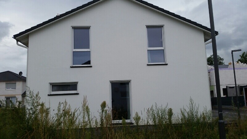 Weißes zweigeschossiges Haus mit drei Fenstern und Tür vorne, überwucherter Vorgarten mit Gras.