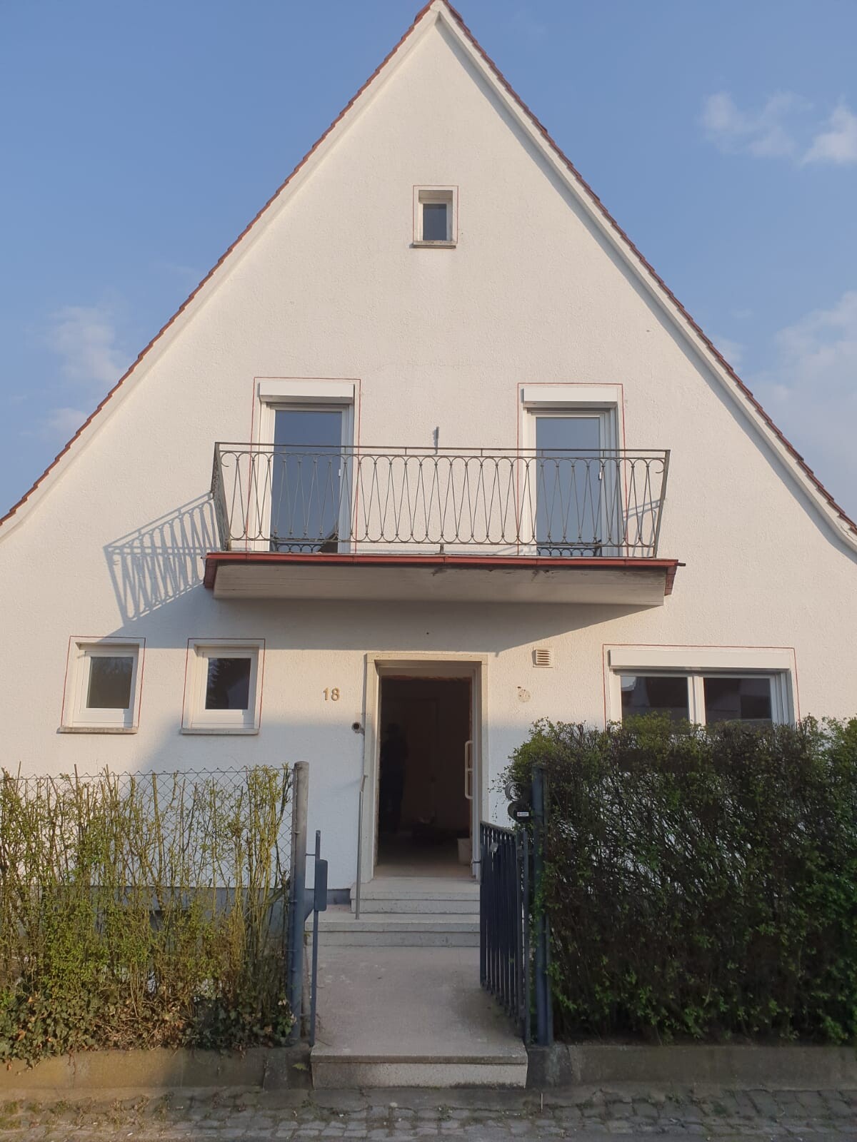 Weißes Haus mit steilem Giebeldach, Balkon und offener Eingang; Hecke im Vordergrund.