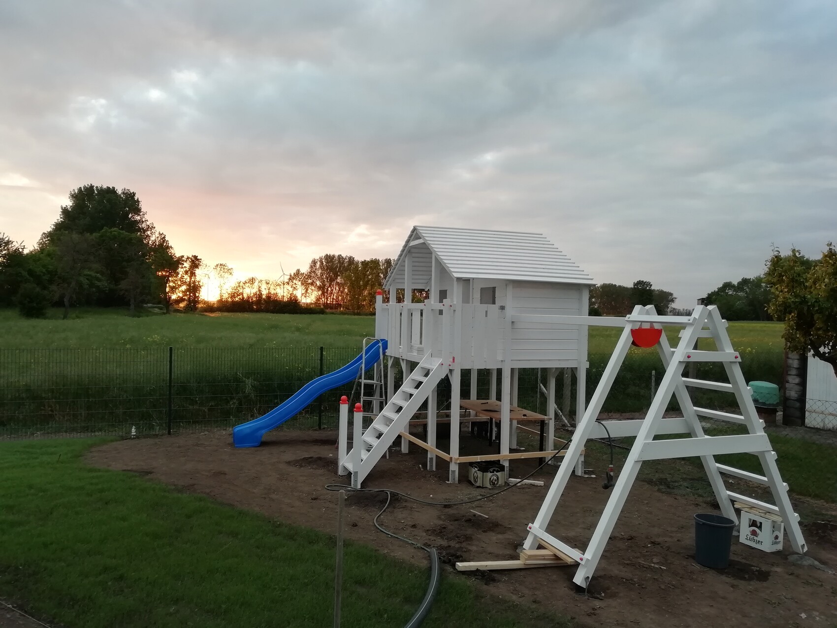Weißes Spielhaus auf Stelzen mit blauer Rutsche im Garten bei Sonnenuntergang.