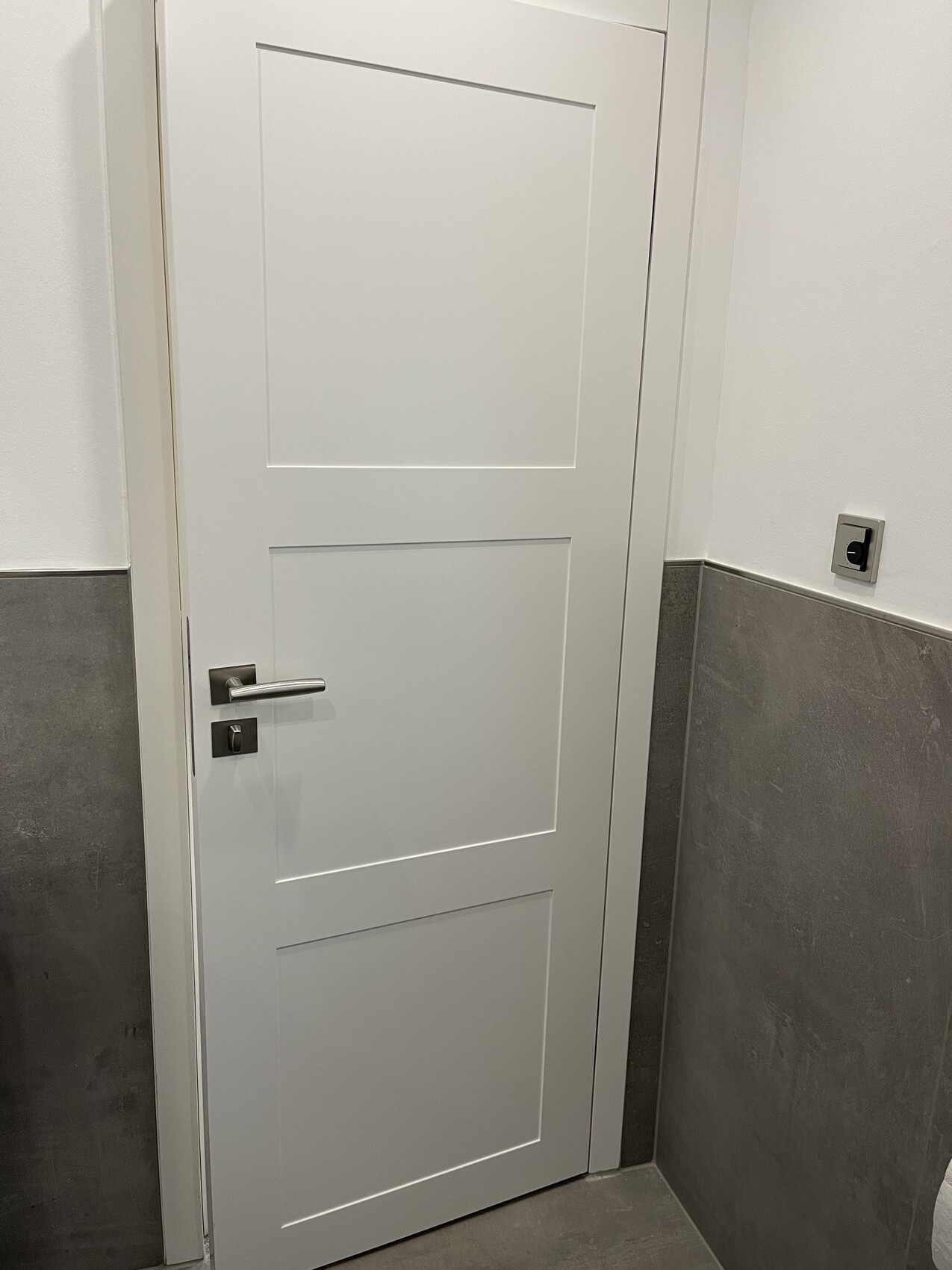 Weiße Tür mit drei Paneelfeldern, Metallgriff und Schloss; graue Fliesenwand daneben.